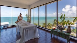 Un cuarto de spa con camilla de masaje, amplias ventanas con vista al océano y piso de madera.