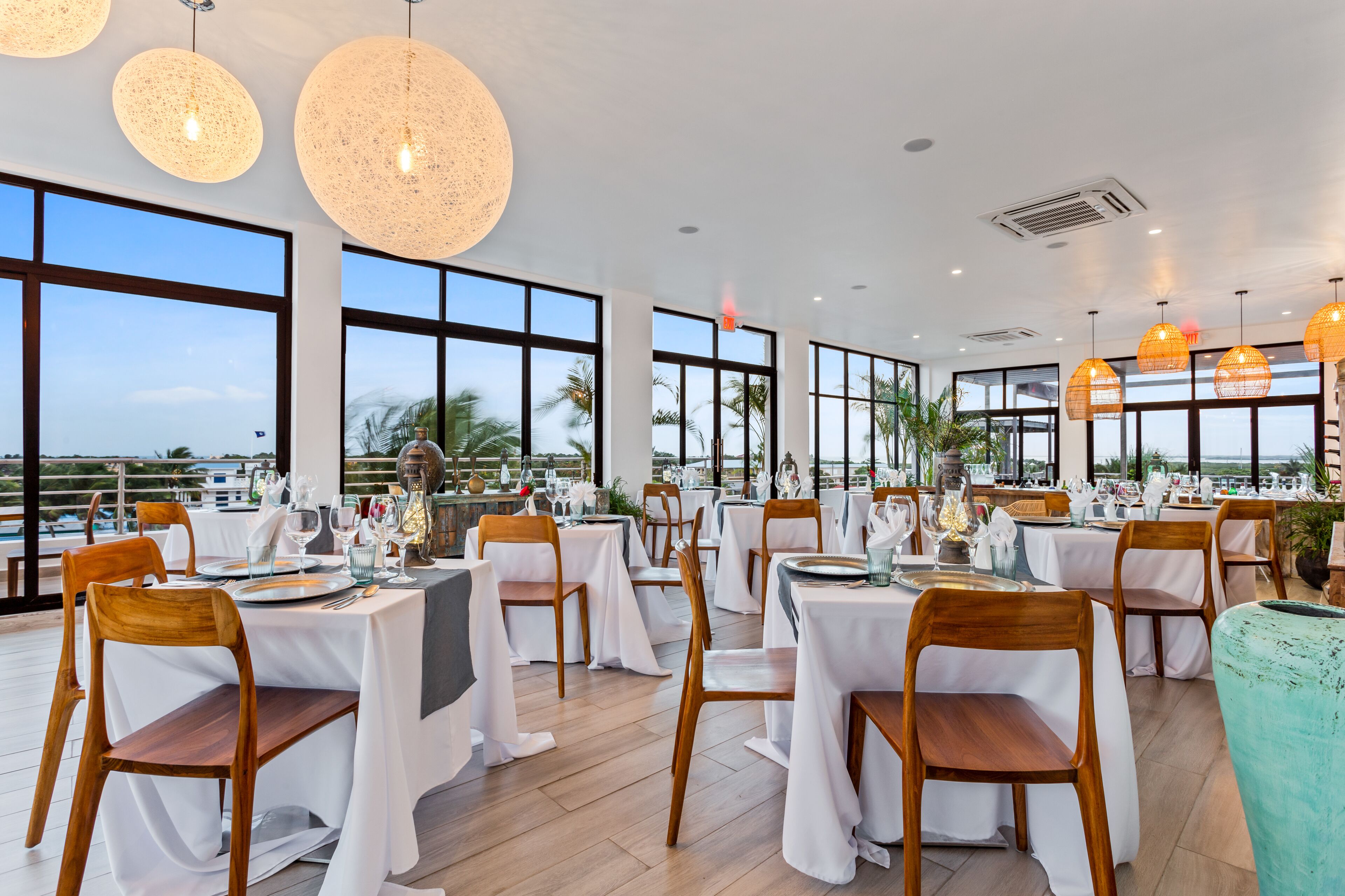 A restaurant with multiple tables set for dining, large windows, and hanging light fixtures.