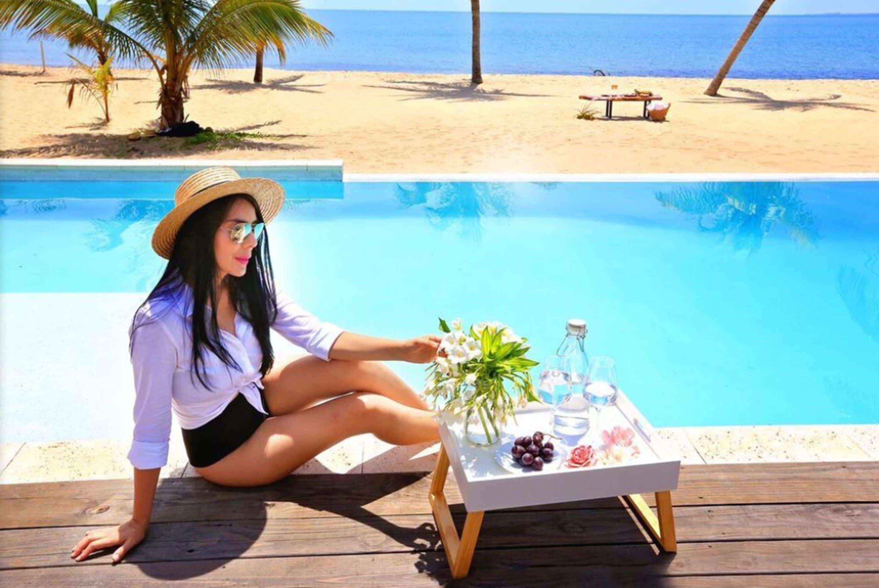 Una persona sentada junto a una mesa de piscina, con una bebida, flores y aperitivos.