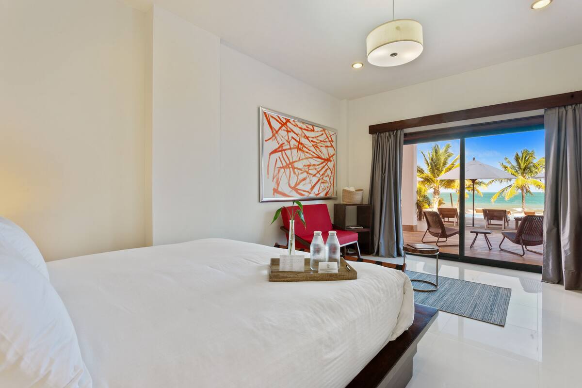 Une chambre d’hôtel avec un grand lit, un bureau avec une chaise, une vue sur la plage et un cadre tropical.