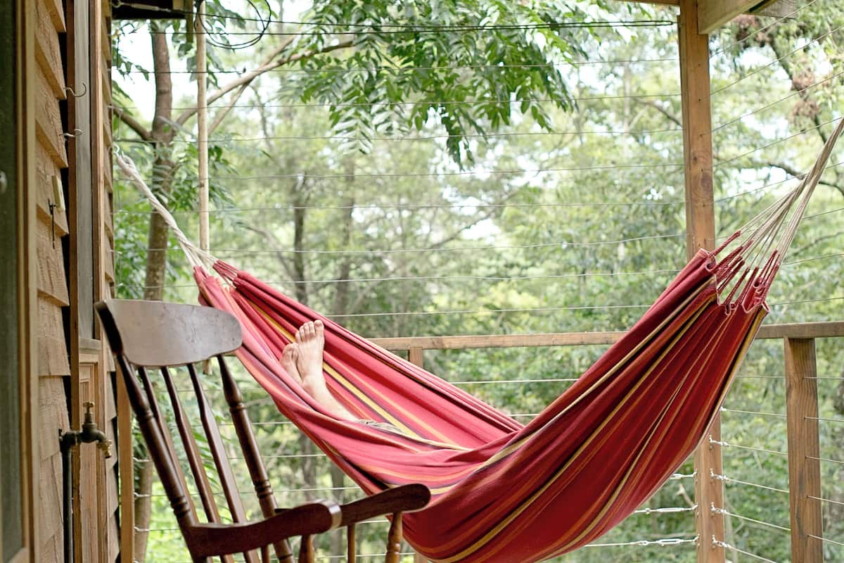 A wooden rocking chair with a red hammock on a wooden deck with a view of trees.