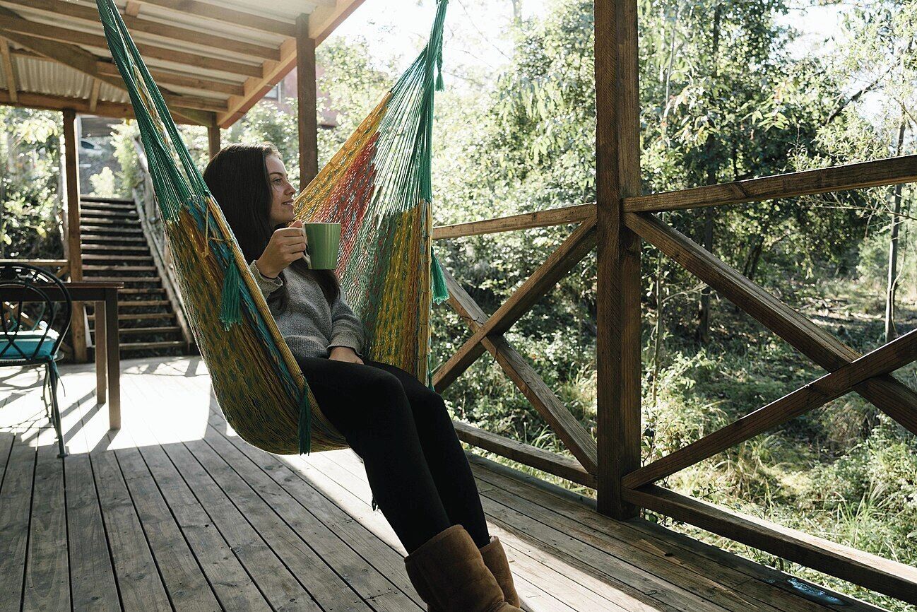 Una persona sentada en una hamaca sobre una terraza de madera, con vista a un área boscosa.