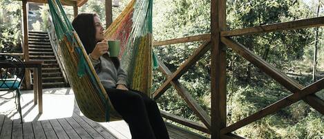 Una persona sentada en una hamaca sobre una terraza de madera, con vista a un área boscosa.