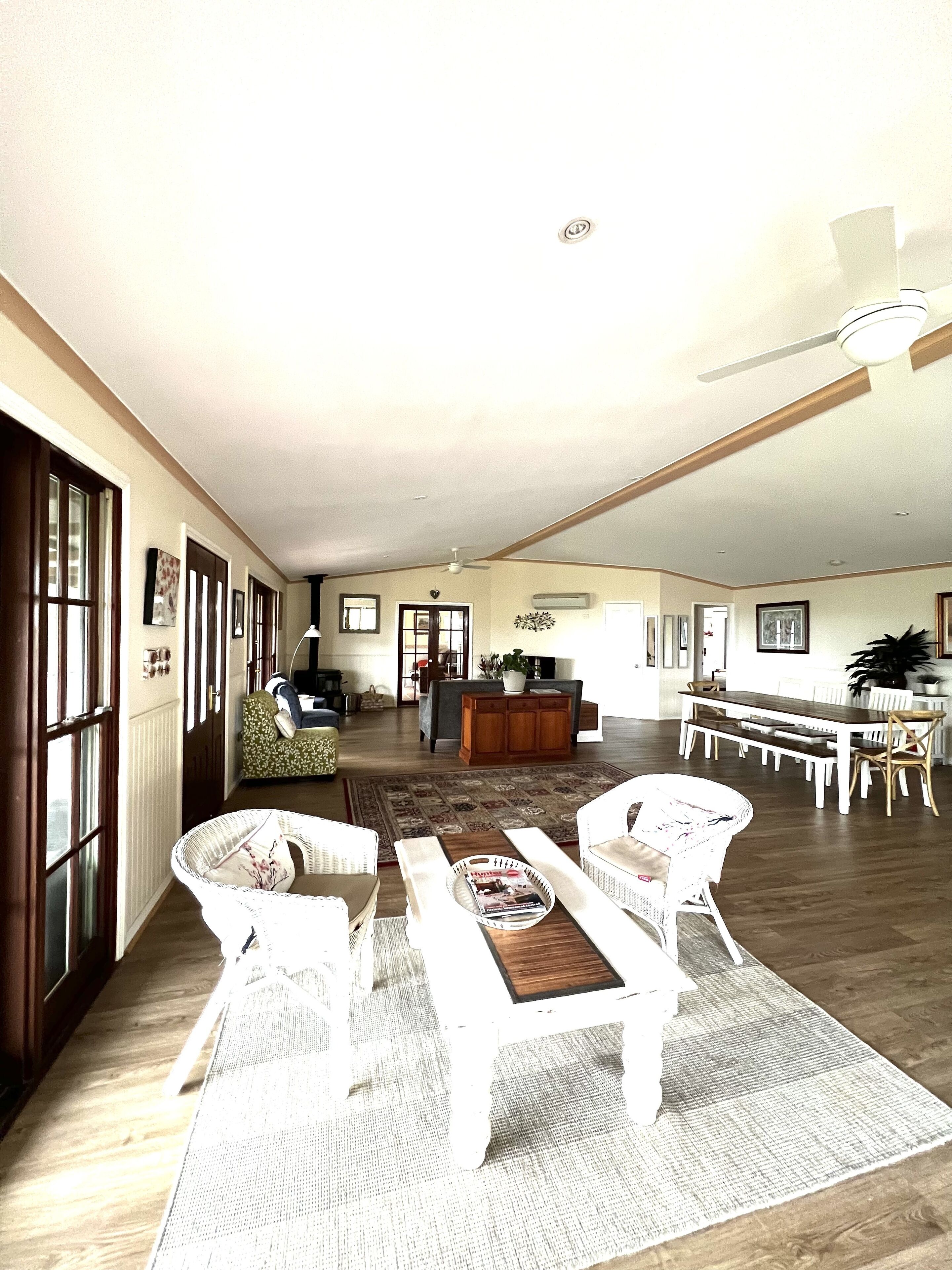 A spacious living room with a dining area, featuring a large wooden dining table, white wicker chairs, and a ceiling fan.