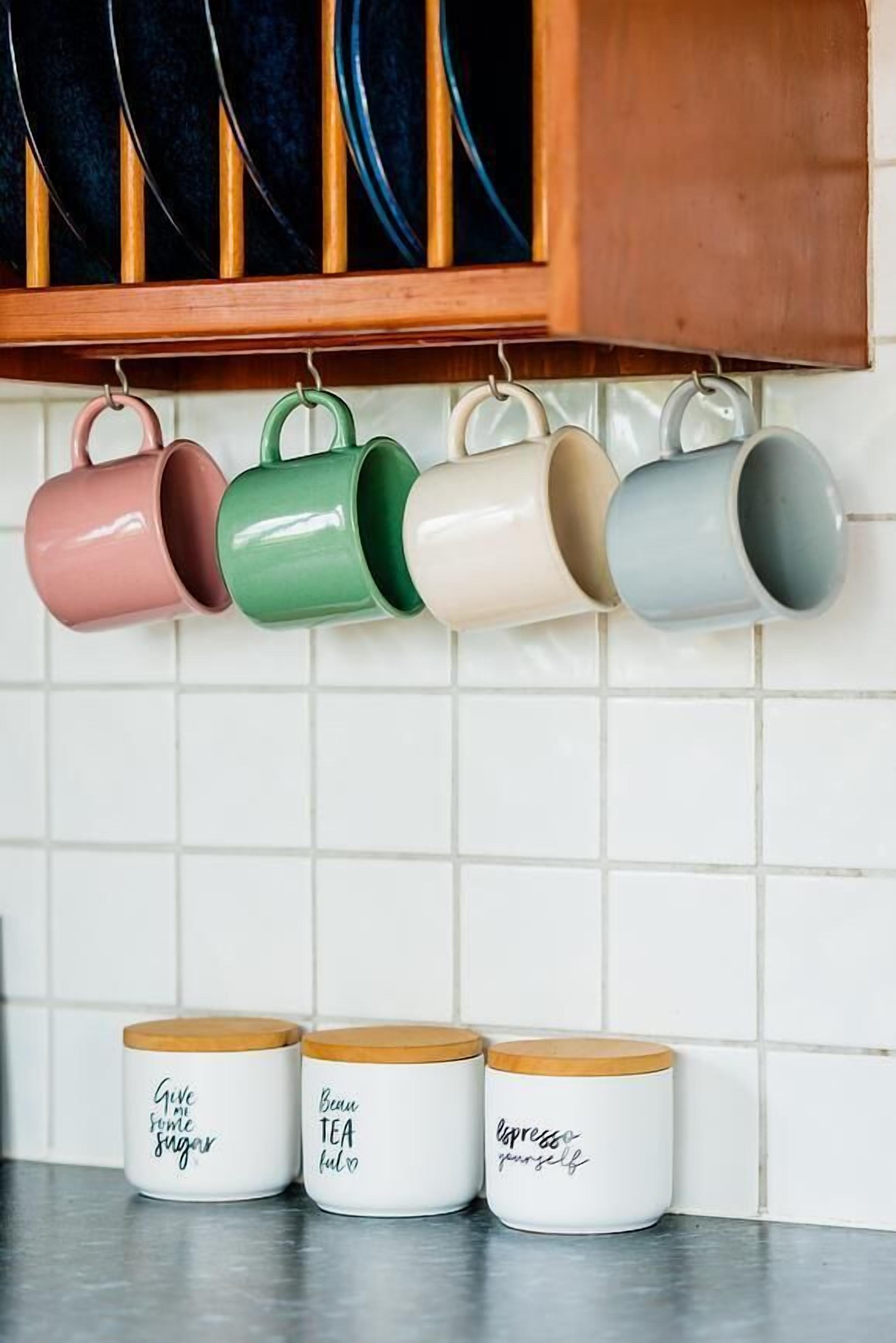 A kitchen with a hanging rack of colorful mugs and three white containers with wooden lids on the counter.