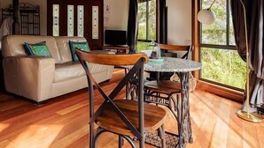 A cozy room with a wooden ceiling, a sofa with cushions, a dining table with chairs, and large windows offering a view of greenery.