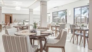 Een ruime eetruimte met ronde tafels en gestreepte stoelen, grote ramen met uitzicht op palmbomen, en een licht, modern interieur.