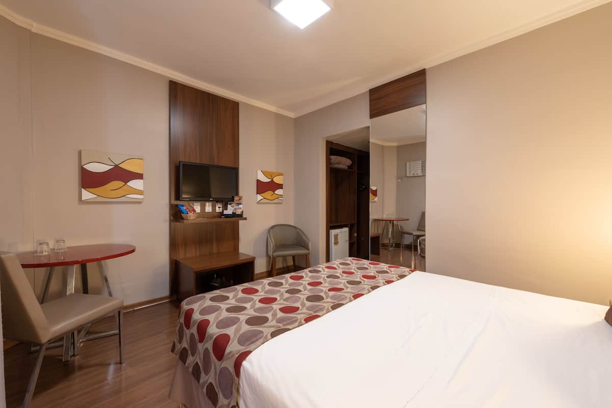 Una habitación de hotel con una cama, un escritorio, una silla, un televisor y obras de arte en las paredes.