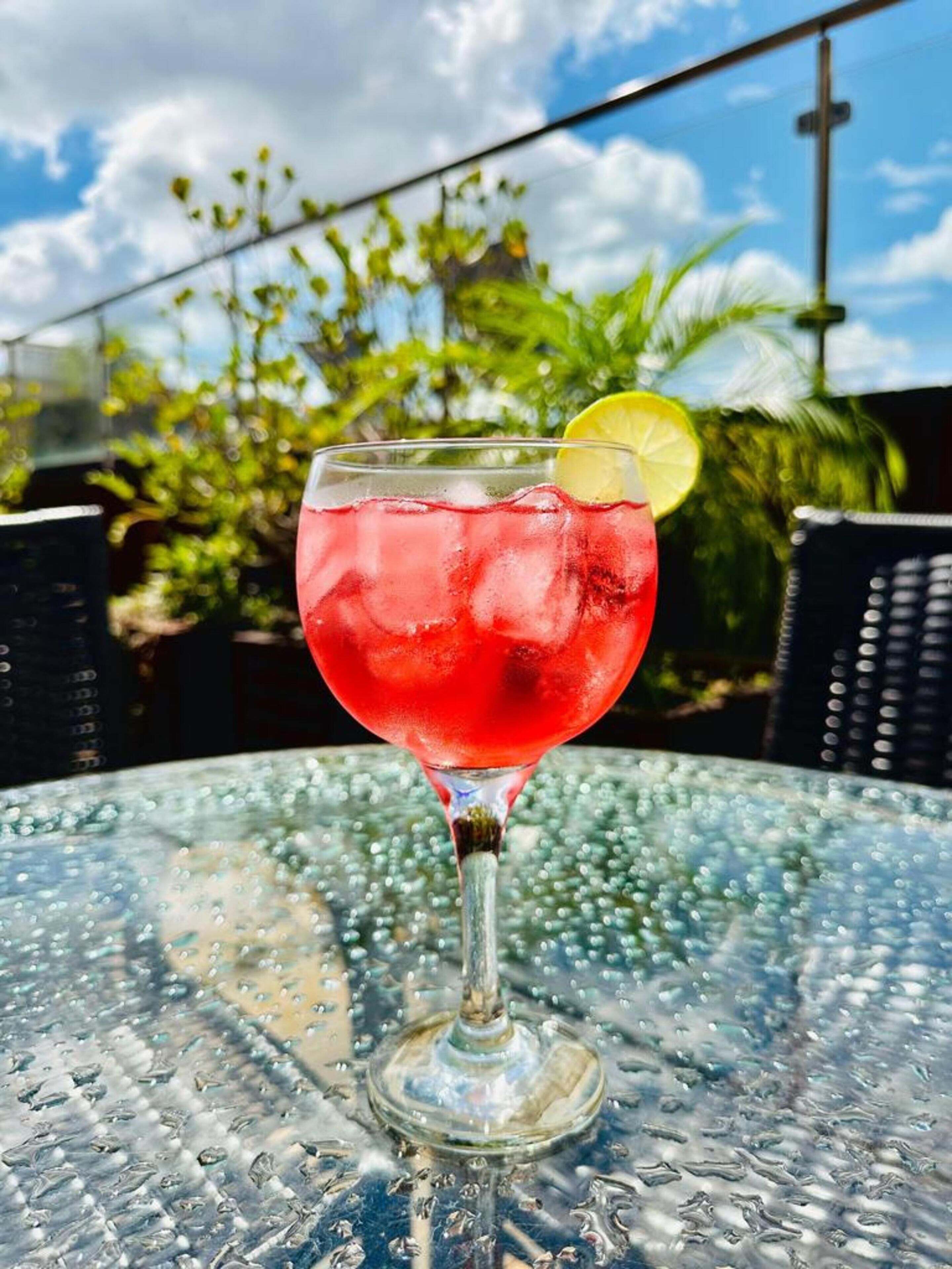 A glass of red drink with a lemon slice on a glass table outdoors.