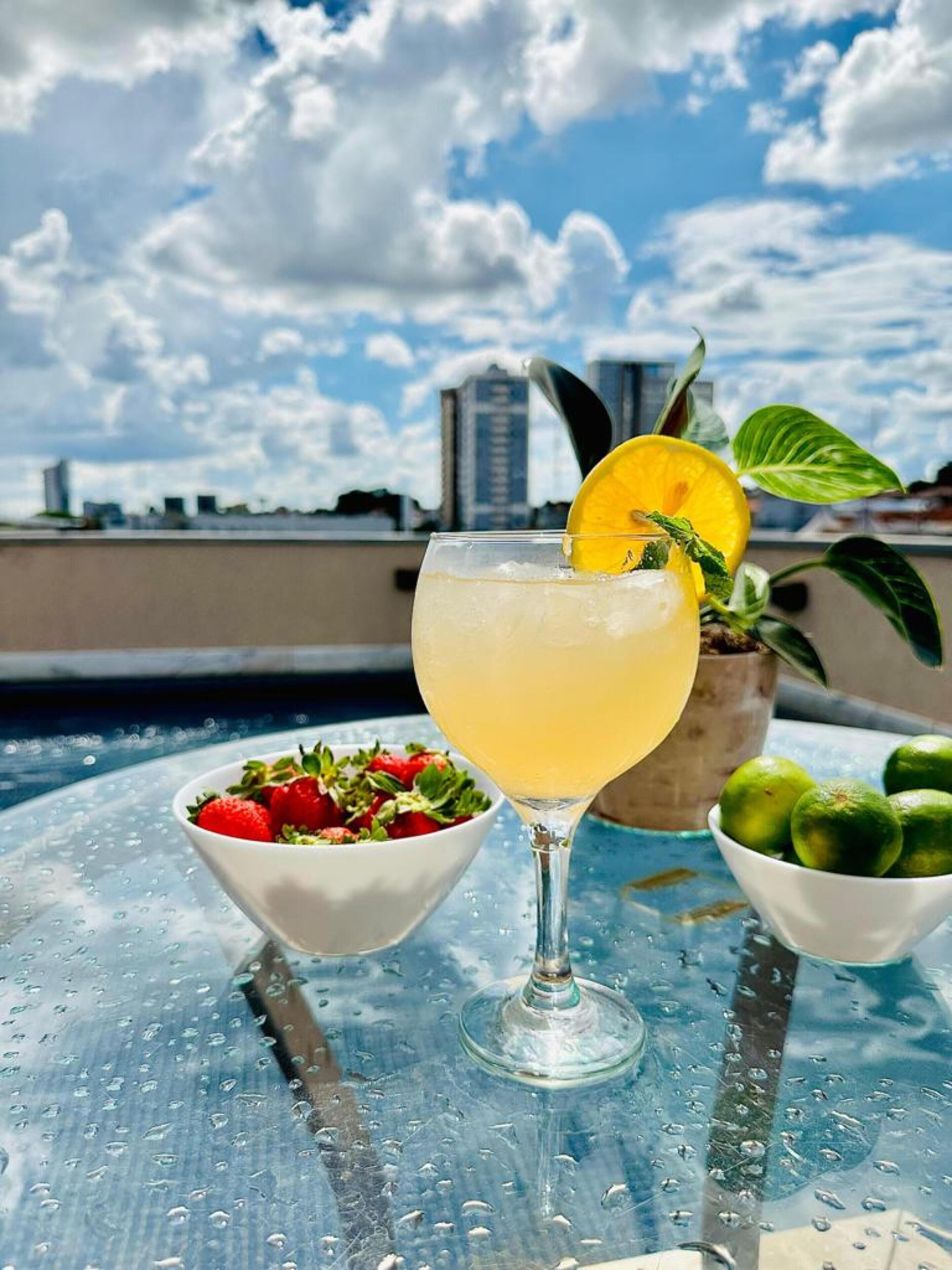 A cocktail glass with a lemon slice garnish, a bowl of strawberries, and a bowl of limes on a table with a cityscape view in the background.