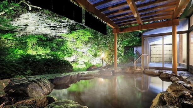 Een traditioneel Japans onsen met een houten constructie, een stenen bad omringd door rotsen en een weelderige groene achtergrond.
