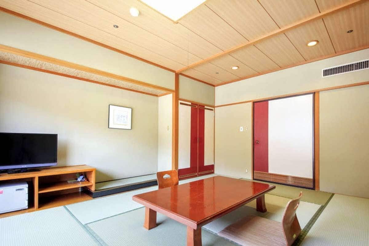 Ein traditioneller japanischer Raum mit Tatami-Böden, einem niedrigen Holztisch, einem Fernseher auf einem Regal und Schiebetüren.