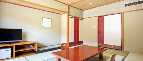 Ein traditioneller japanischer Raum mit Tatami-Böden, einem niedrigen Holztisch, einem Fernseher auf einem Regal und Schiebetüren.