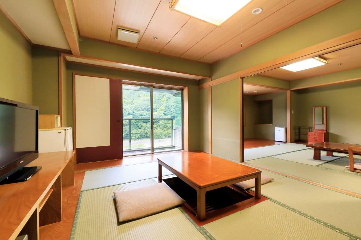 Ein traditioneller japanischer Raum mit Tatami-Boden, einem niedrigen Holztisch, einem Fernseher und Schiebetüren, die auf einen Balkon führen.