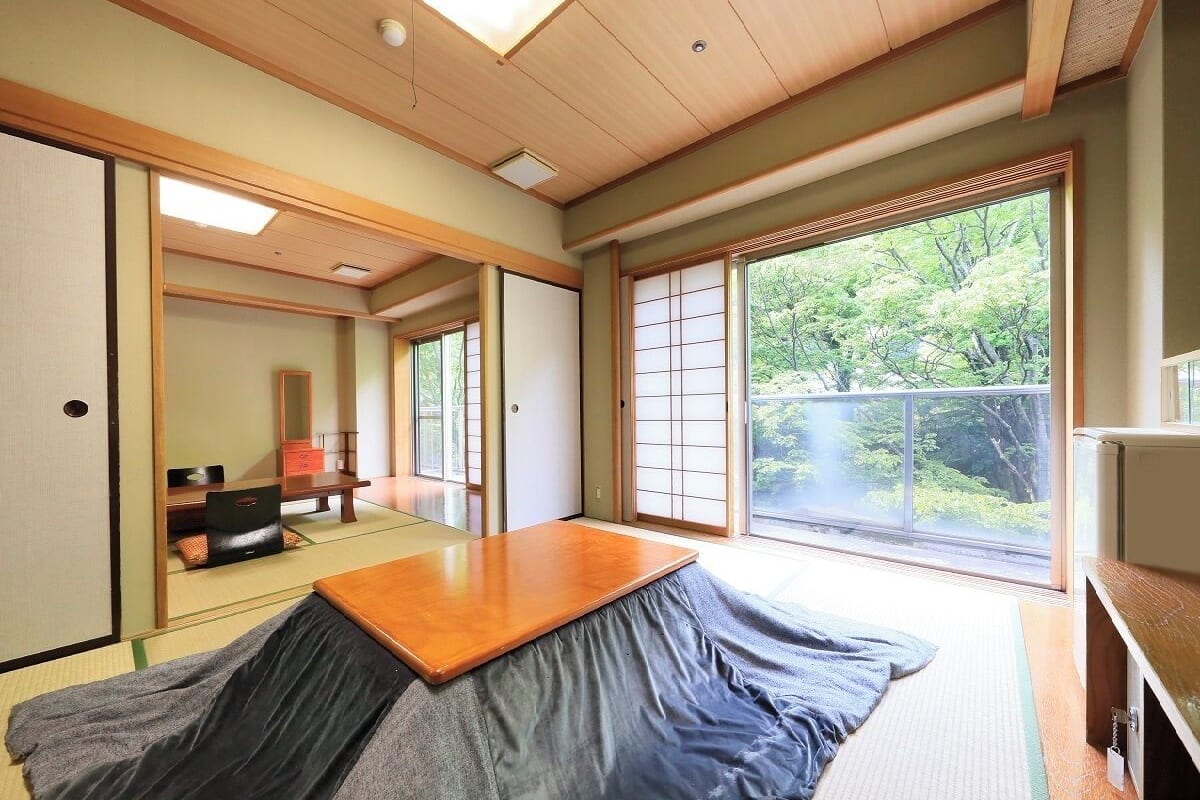 Ein traditioneller japanischer Raum mit Tatami-Bodenbelag, einem Futon, Schiebetüren, einem Holztisch und Blick auf Grünfläche.