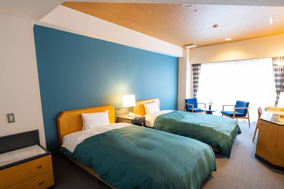 Ein Hotelzimmer mit zwei Betten, einem Schreibtisch, einem Stuhl und einem Fenster mit Vorhängen.