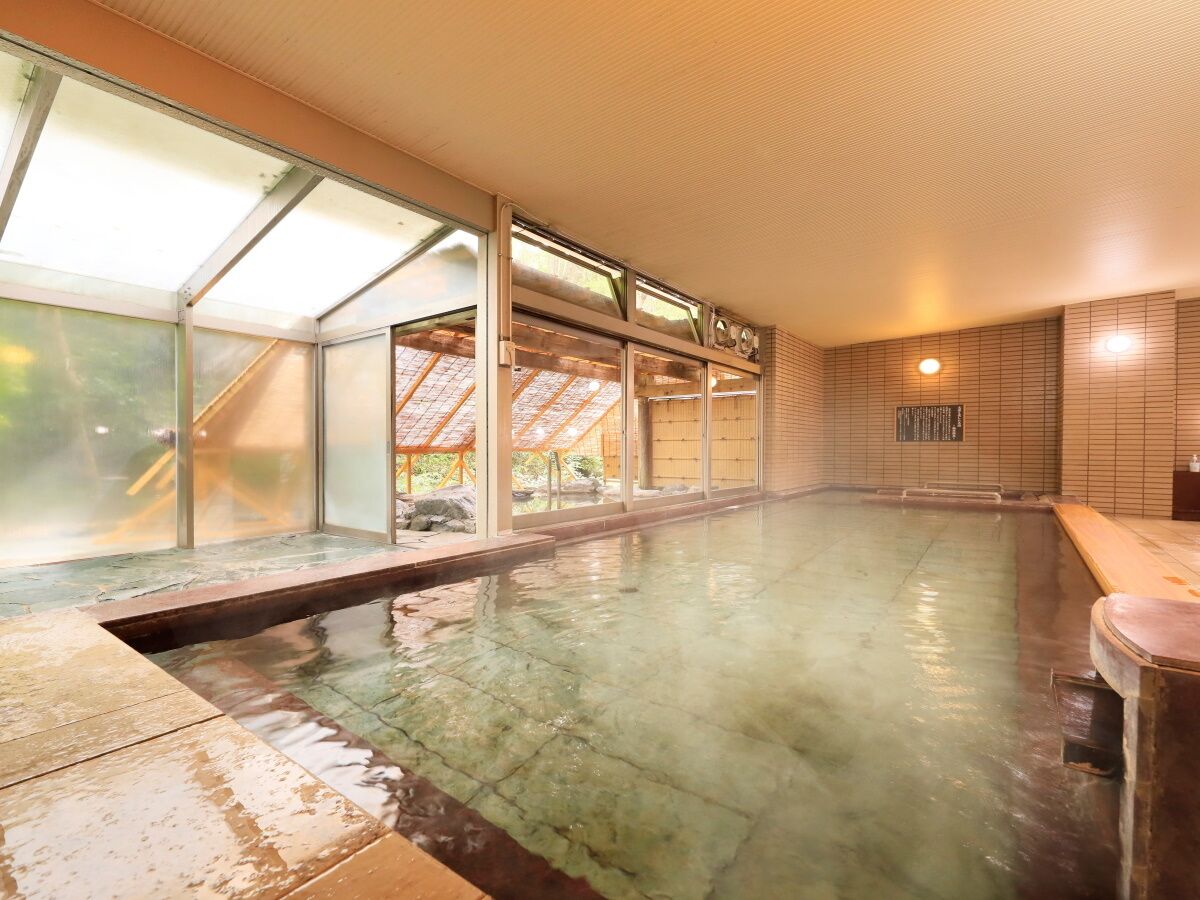 A traditional Japanese onsen with a stone bath, wooden benches, and a view of the outdoors through large glass doors.