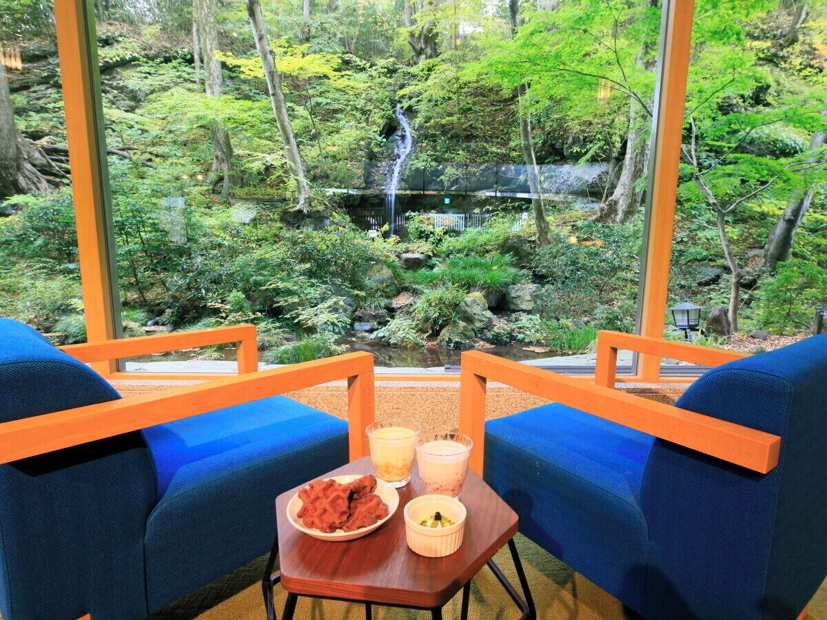 Una habitación con dos sillas azules y una pequeña mesa con comida y bebidas. Se ve una vista de un bosque y una cascada a través de amplios ventanales.