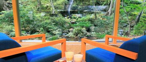 Una habitación con dos sillas azules y una pequeña mesa con comida y bebidas. Se ve una vista de un bosque y una cascada a través de amplios ventanales.