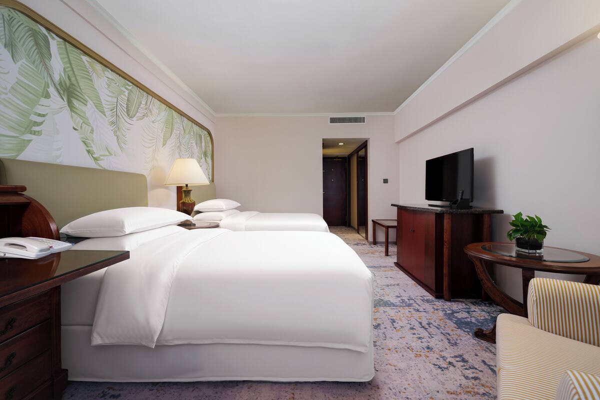 Quarto de hotel com cama, mesa de cabeceira, luminária, TV e pequena escrivaninha com cadeira.