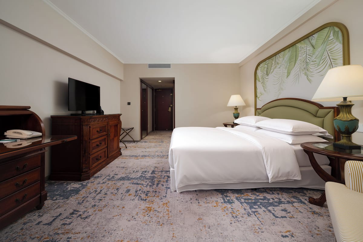 Quarto de hotel com uma cama, televisão, escrivaninha com telefone e um quadro na parede.