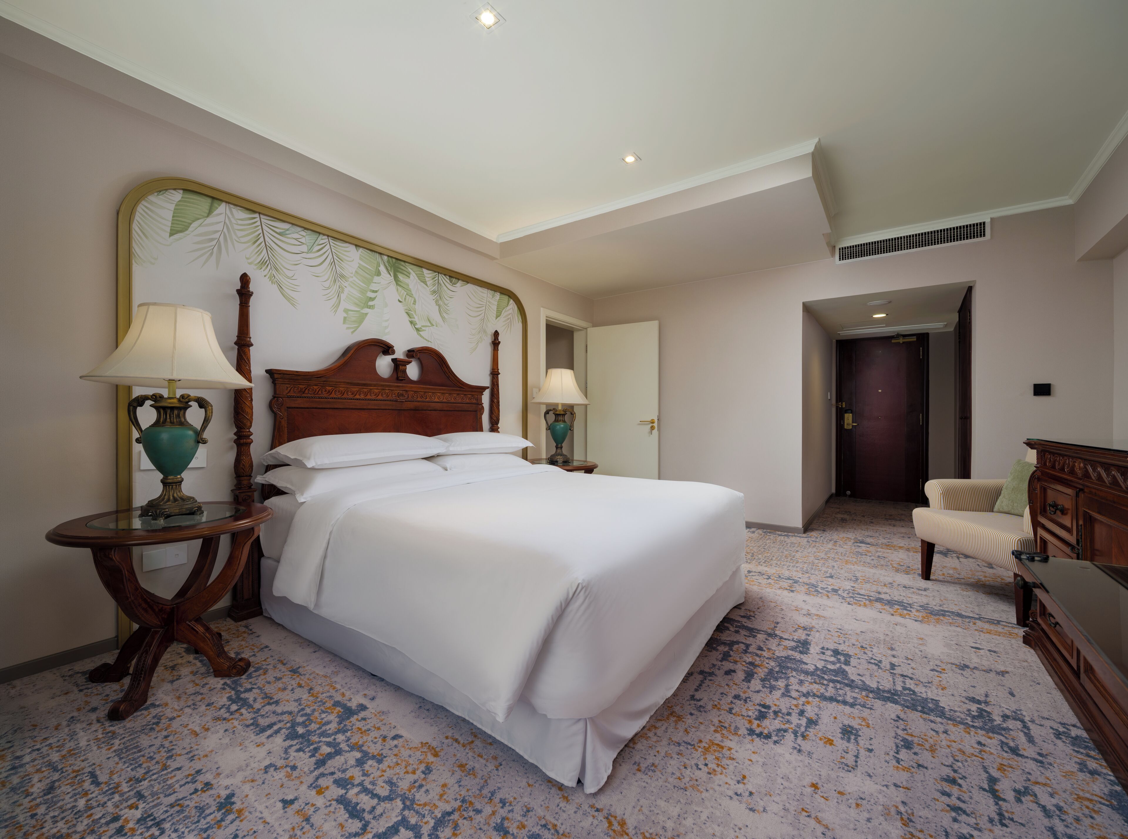 Une chambre d’hôtel avec un grand lit, deux tables de chevet avec des lampes, une tête de lit en bois, un miroir, une chaise et un sol recouvert de moquette.