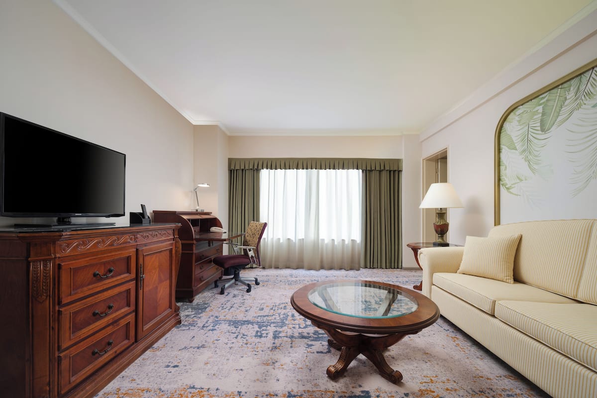 Quarto de hotel com televisão de tela plana, cómoda de madeira, mesa de centro de vidro, sofá listrado e uma grande janela com cortinas.