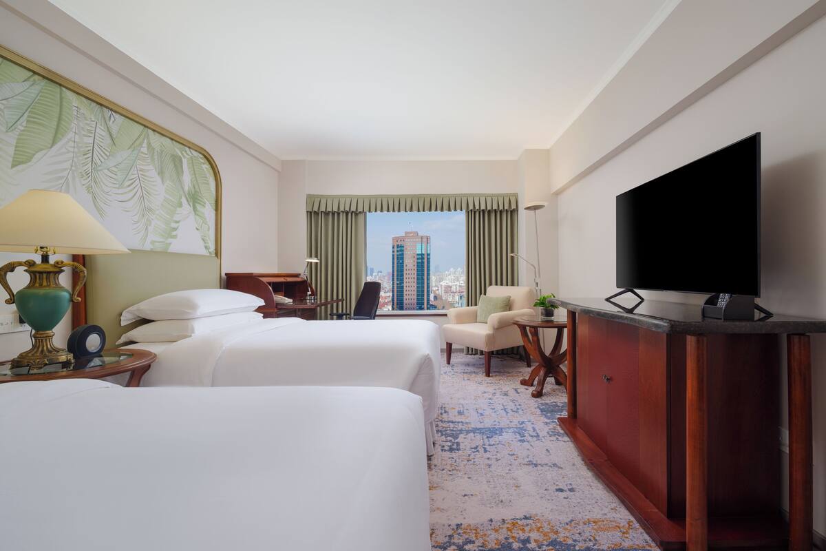 Quarto de hotel com duas camas, televisão, escrivaninha, cadeira e vista para a paisagem urbana.