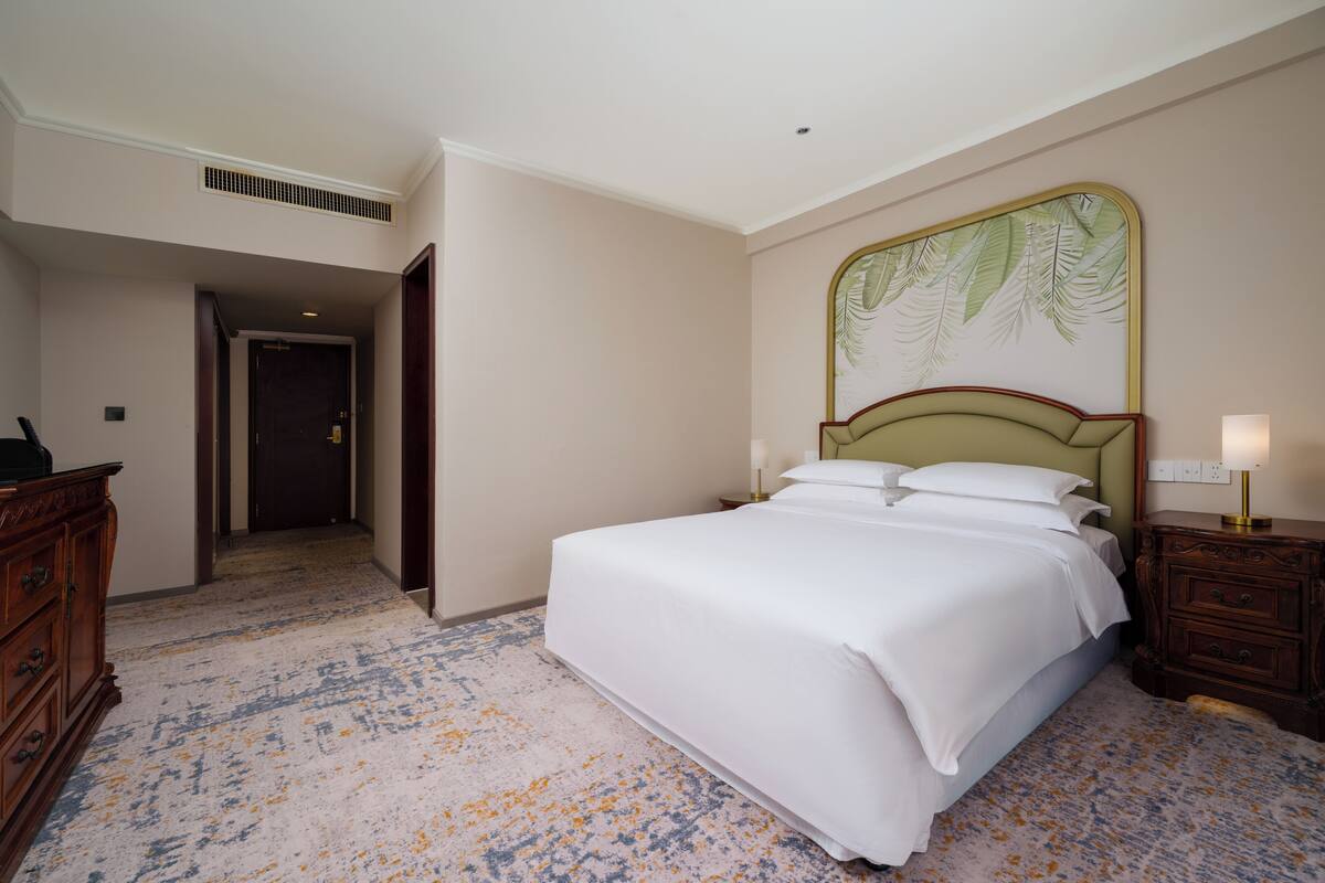 Quarto de hotel com uma cama, mesa de cabeceira, penteadeira, porta que dá para outro cômodo e parede com padrão de folhas.