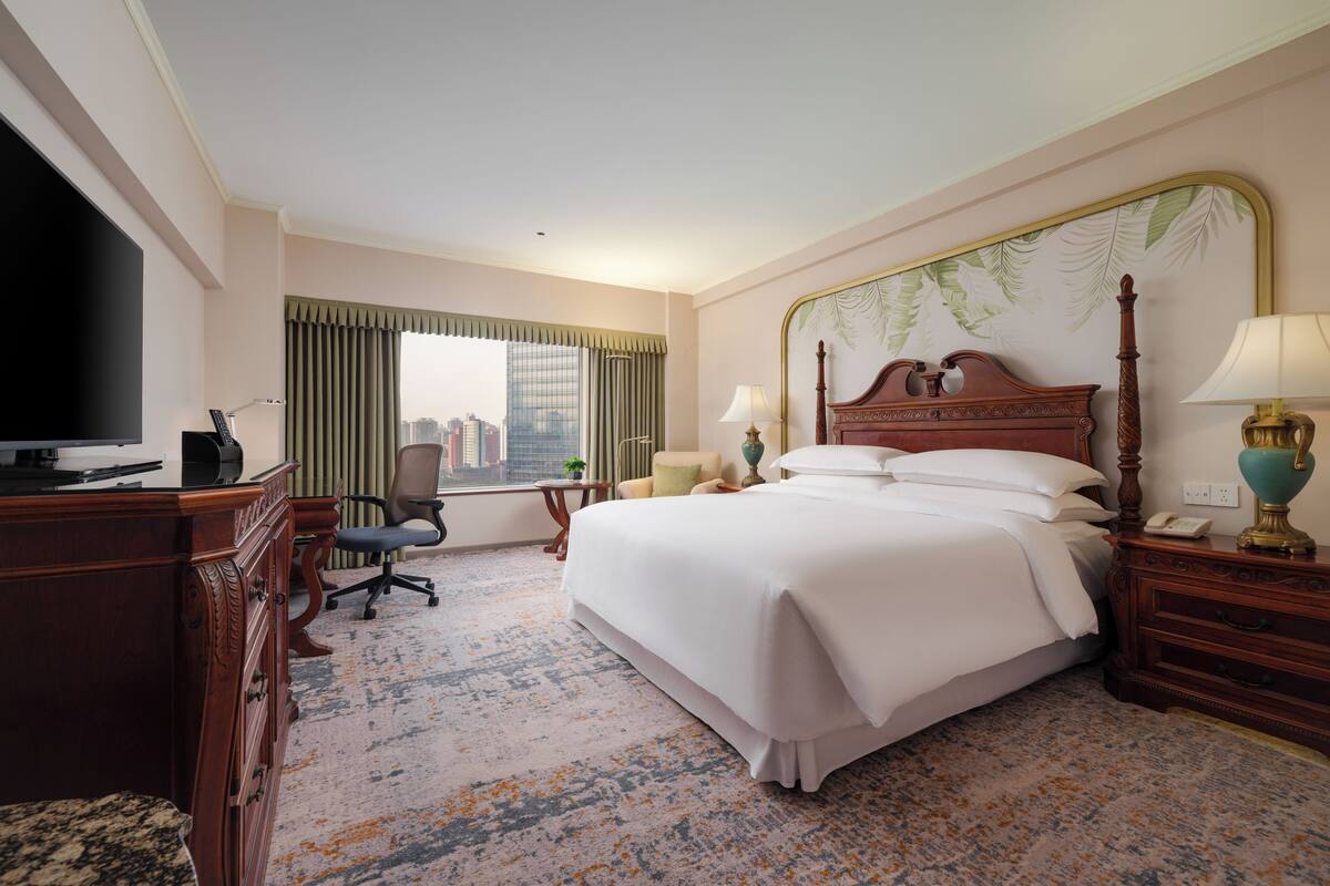Quarto de hotel com cama, secretária com televisão, poltrona, mesa de cabeceira com abajur e vista para a paisagem urbana pela janela.