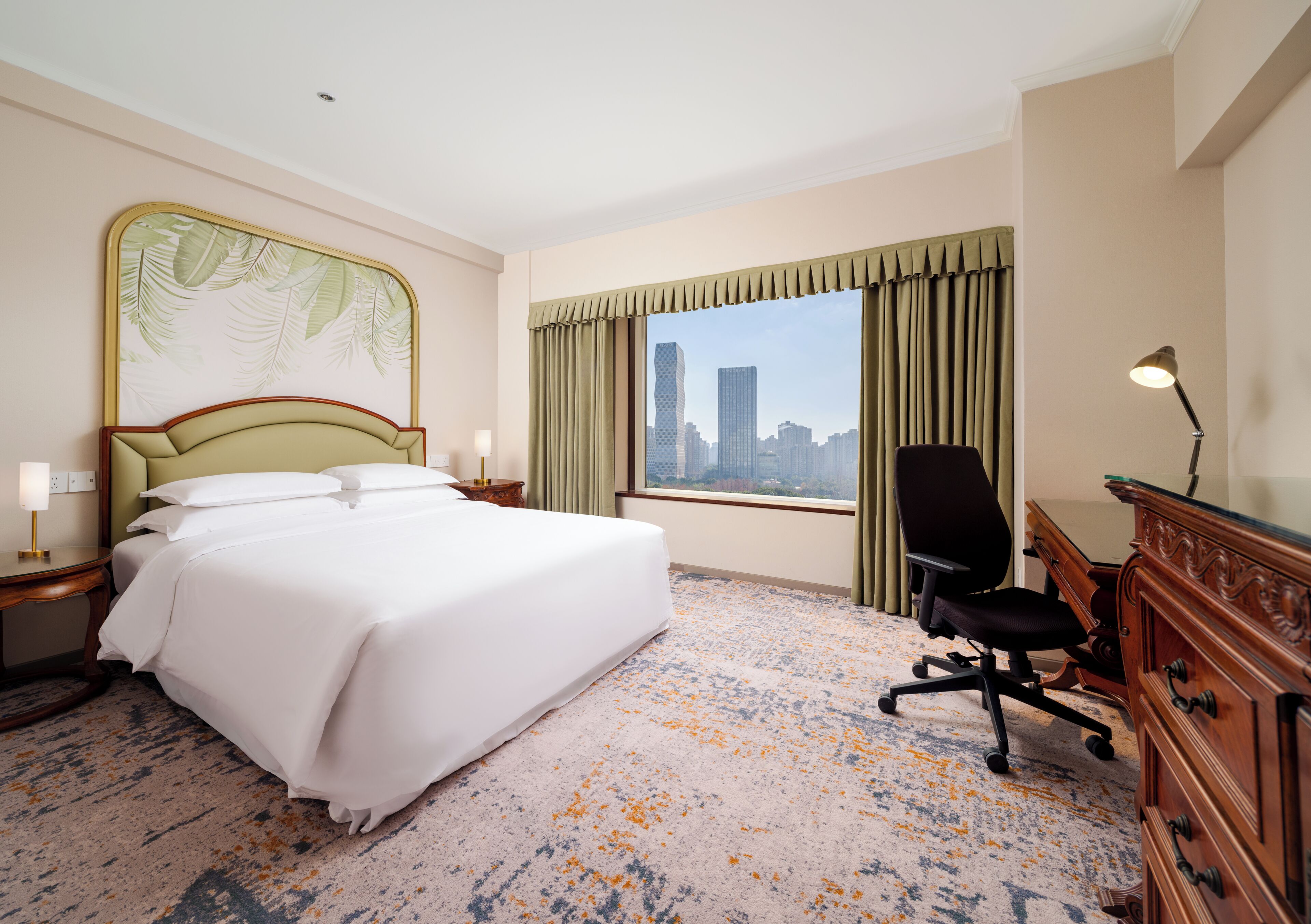 Hotelkamer met een groot bed, een bureau, een stoel, een lamp en uitzicht op een stadsgezicht door het raam.