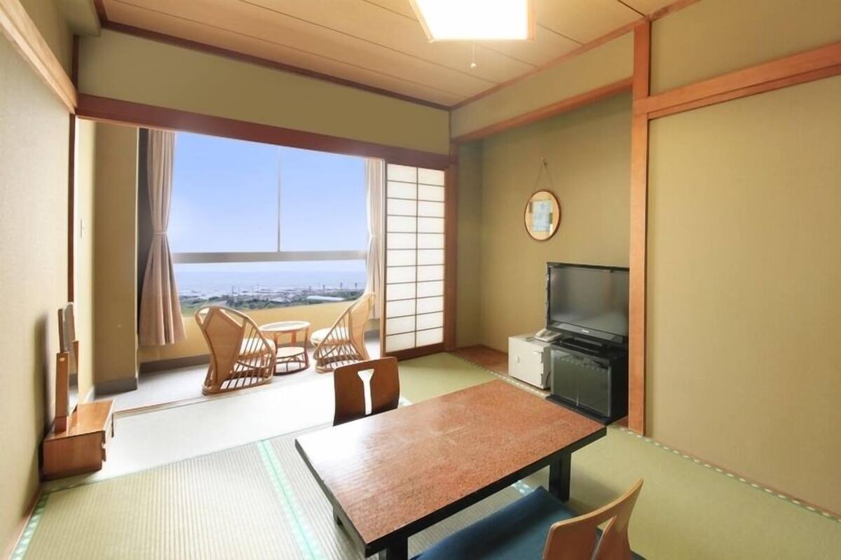 A traditional Japanese room with tatami flooring, a low table, chairs, a TV, and a view of the sea.
