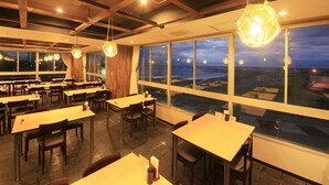 A restaurant with tables and chairs arranged in rows, large windows offering a view of the cityscape at dusk. - Bousou Shirahama Umisato Hotel (Minamiboso)