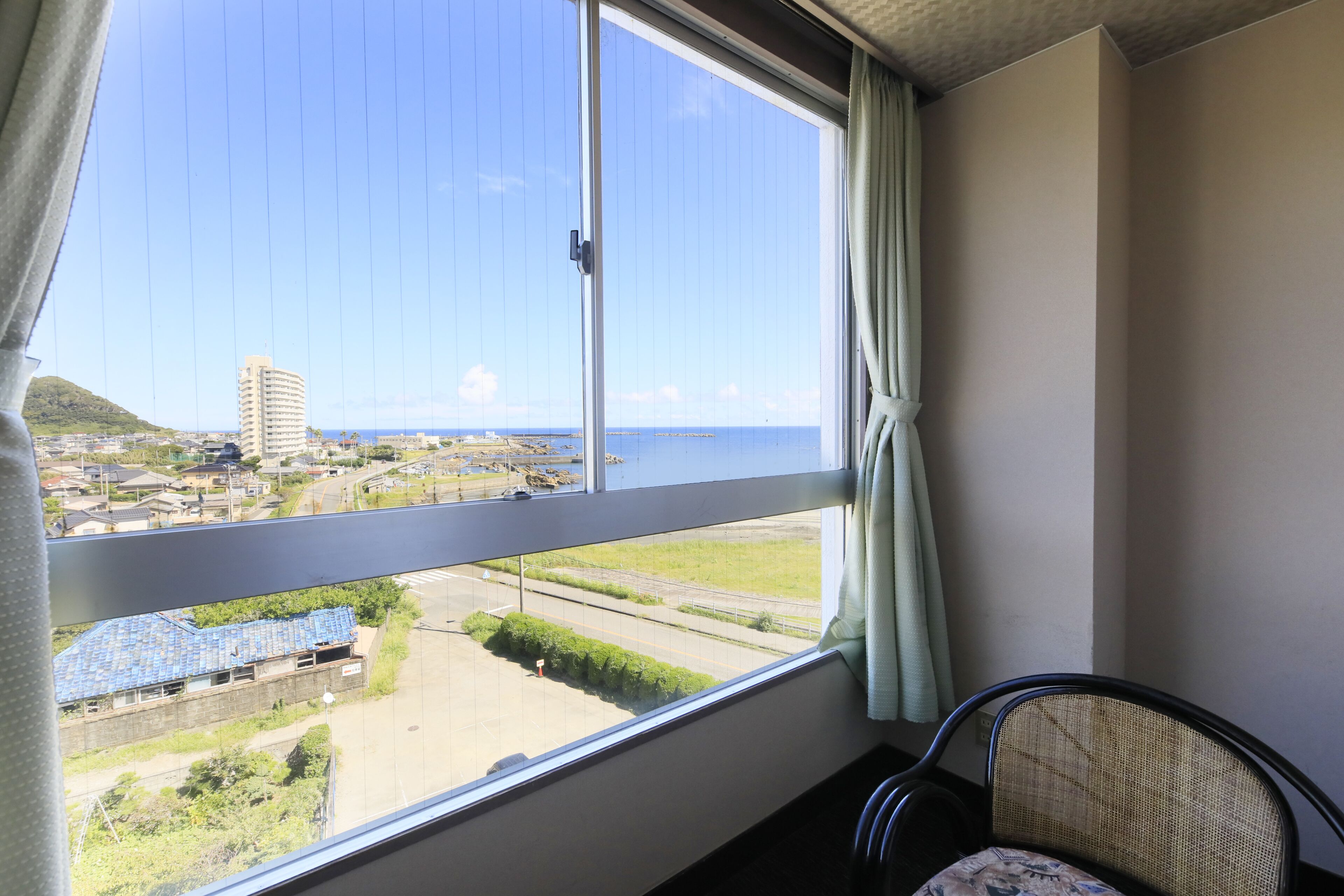 A room with a large window offering a view of a coastal area, a chair, and a curtain.