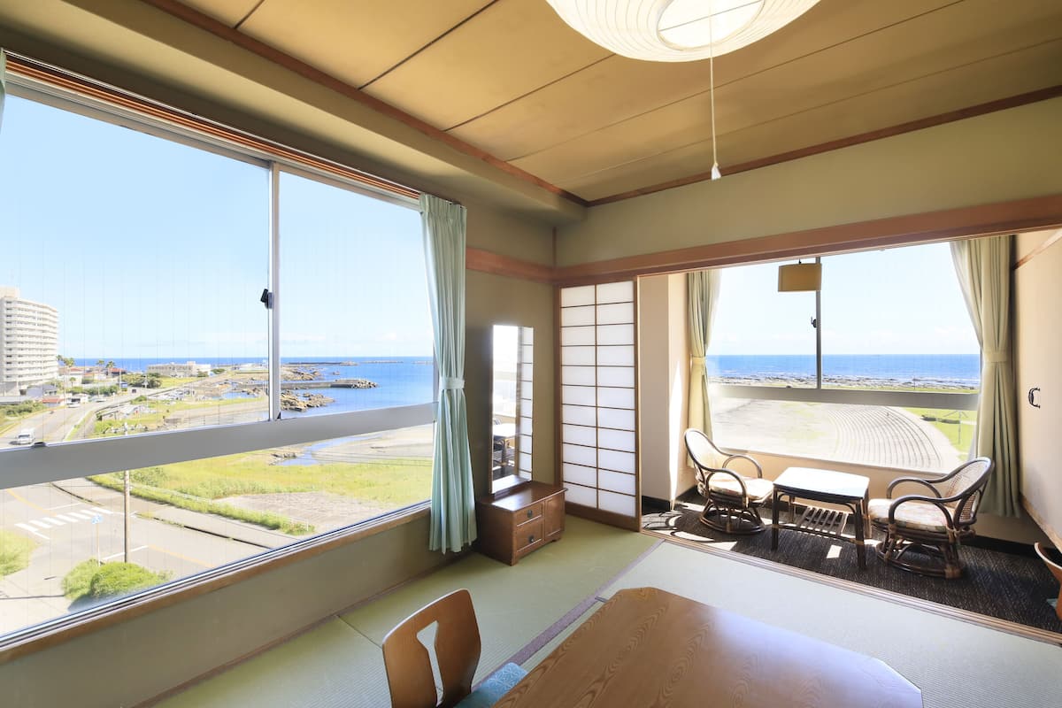 A hotel room with a large window offering a view of the sea and a cityscape. The room includes a bed, a small table, and two chairs.