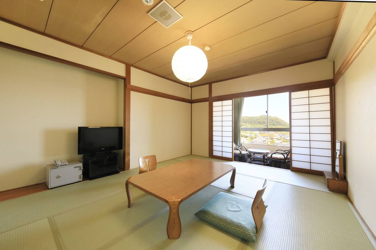 A traditional Japanese room with tatami flooring, a low wooden table, a television, and sliding shoji doors leading to a balcony with a view of a mountain.