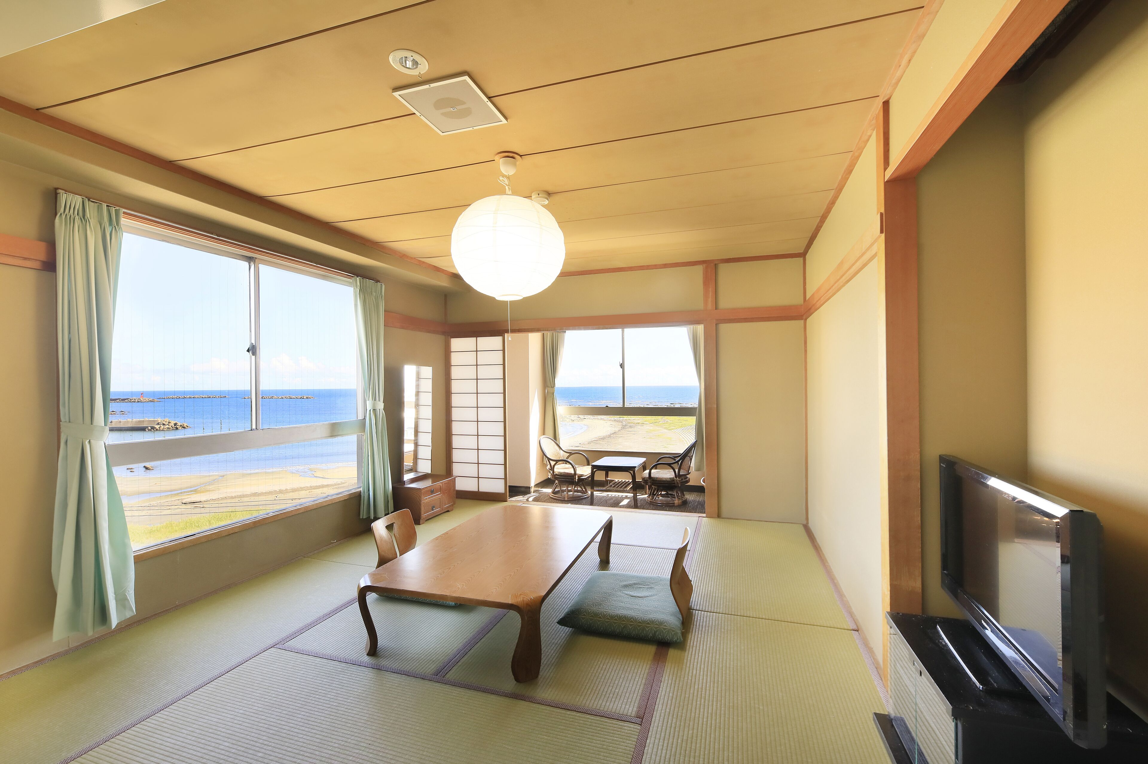 A traditional Japanese room with tatami flooring, a low table, a television, and large windows overlooking a beach.