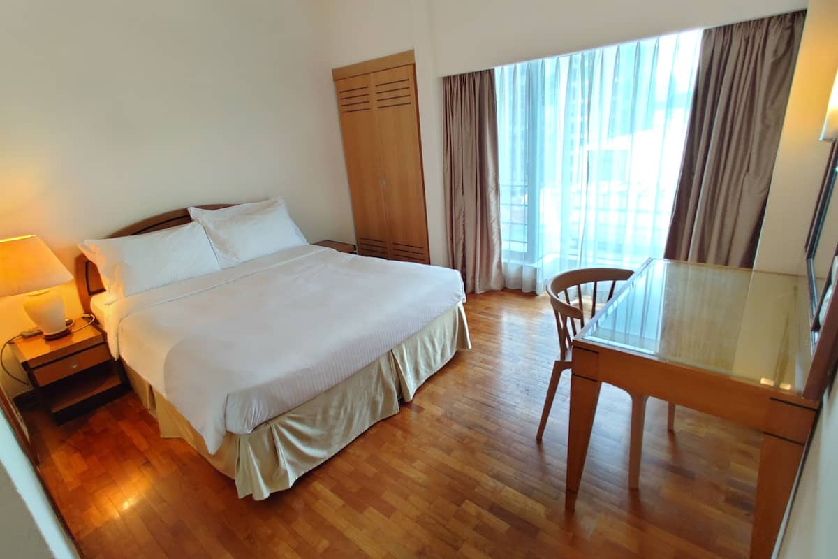 A hotel room with a bed, bedside table, lamp, wooden wardrobe, and a dining table with a chair.