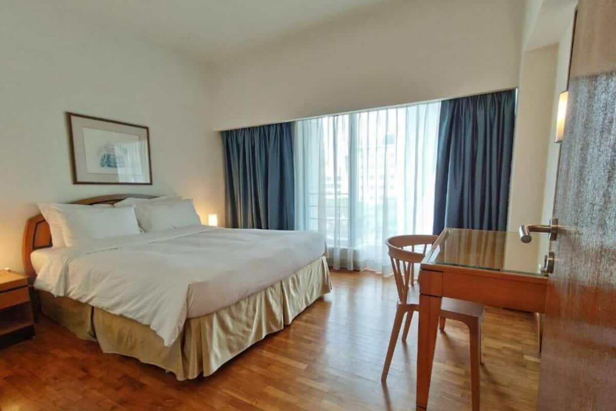 A hotel room with a large bed, a desk, and a chair. There is a window with curtains, a framed picture on the wall, and a door with a handle.