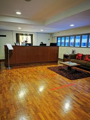 Un área de recepción con un mostrador de madera, dos computadoras, un sofá con cojines rojos, una mesa de centro y una ventana con vistas.