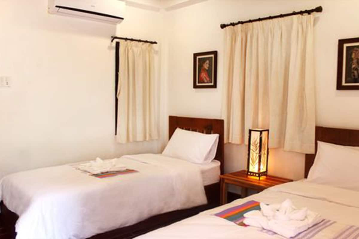 A hotel room with two single beds, a nightstand with a lamp, and a wall-mounted air conditioner.