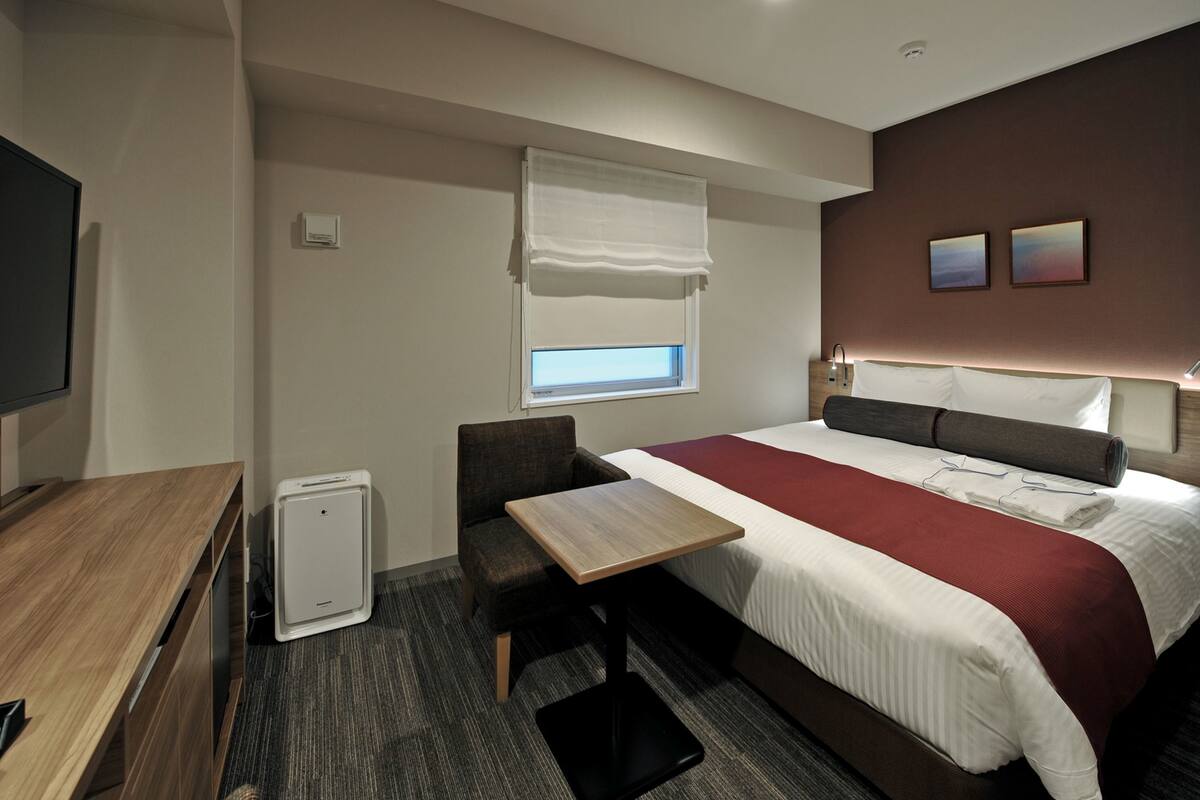 A hotel room with a bed, a desk, a chair, a heater, a window with blinds, and two wall-mounted picture frames.