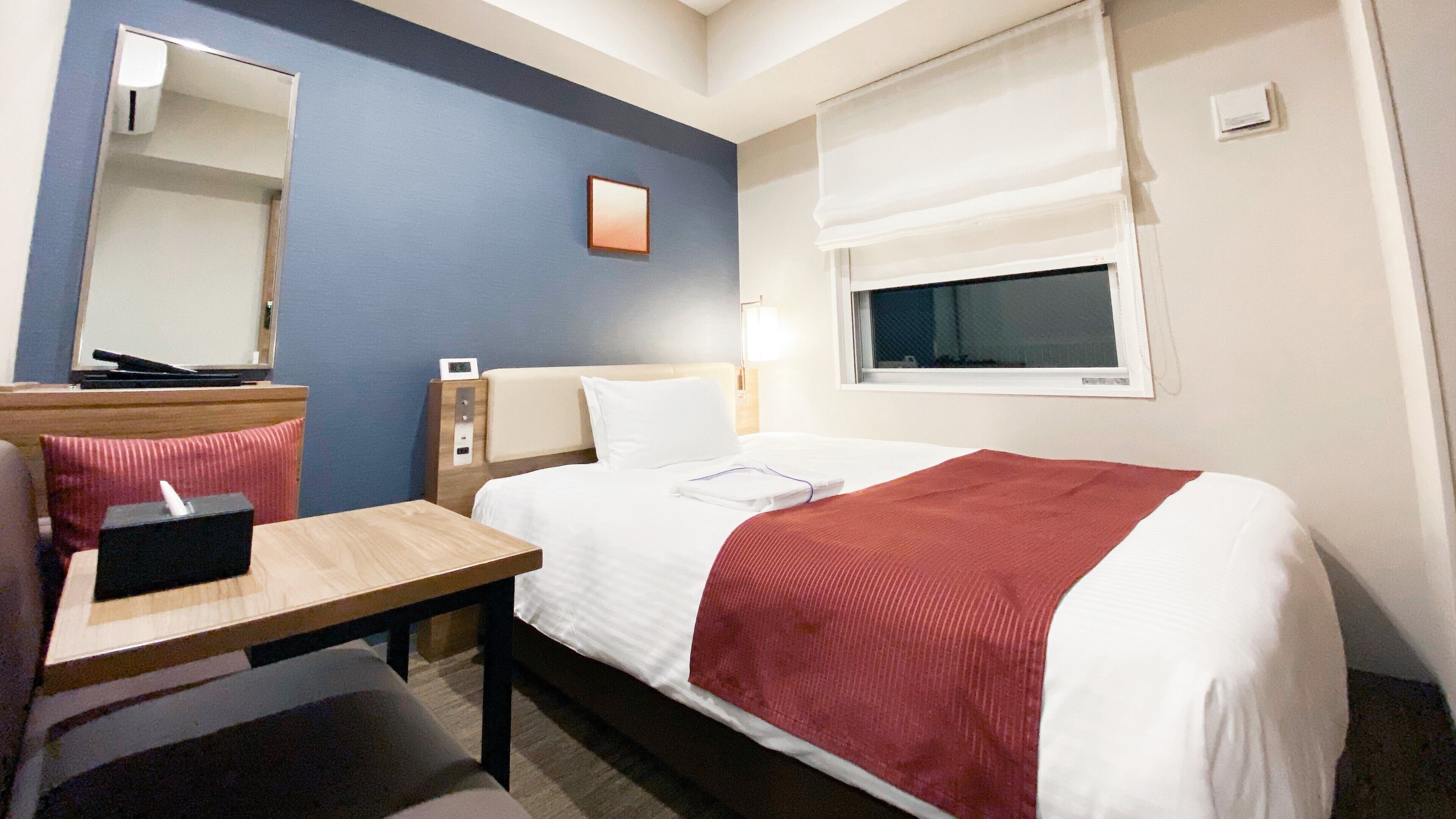 A hotel room with a bed, a desk, a chair, a mirror, a window, and a bedside table.