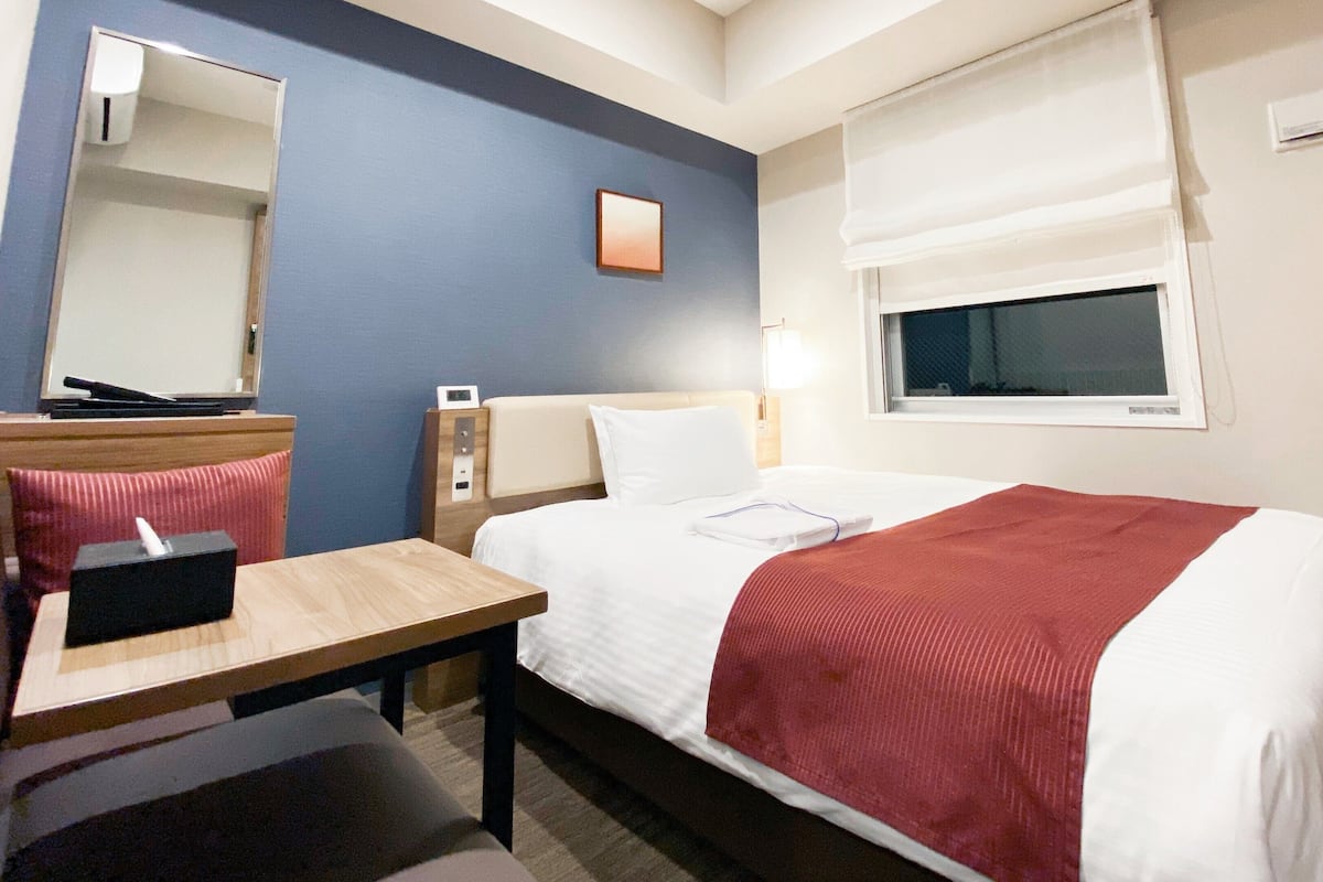A hotel room with a bed, a desk, a chair, a mirror, a window, and a bedside table.
