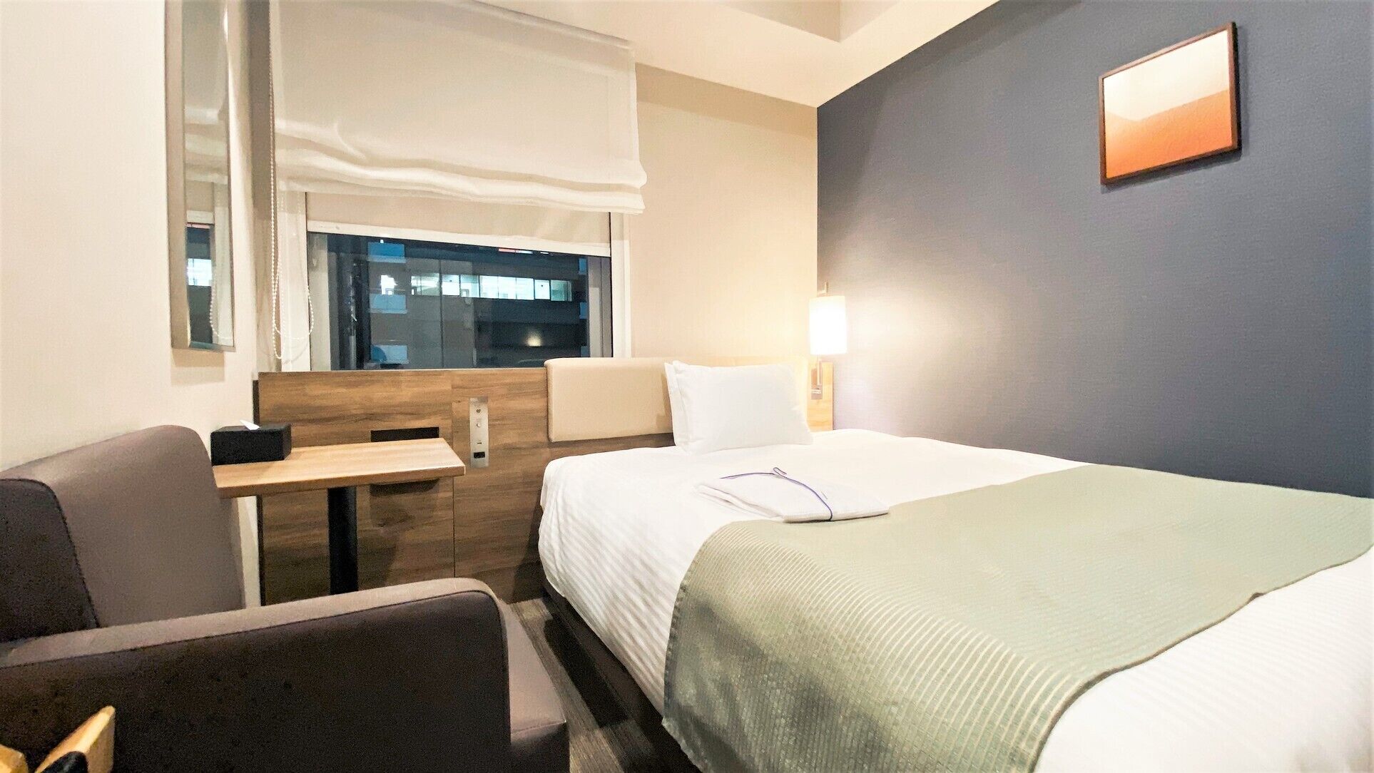 Une chambre d’hôtel avec un lit, un bureau, une chaise, une fenêtre et un tableau accroché au mur.