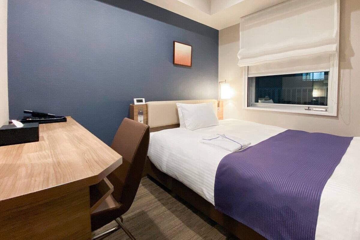 A hotel room with a bed, bedside table, chair, lamp, and a window with a view.