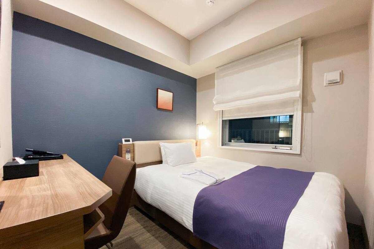A hotel room with a bed, a desk, a chair, a bedside table, a lamp, and a window with a white blind.