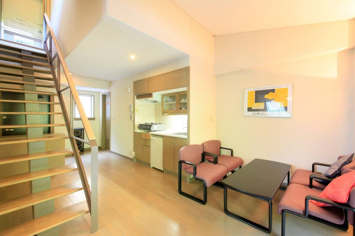 A modern apartment with a wooden staircase, a dining area with pink chairs, and a kitchen with wooden cabinets.