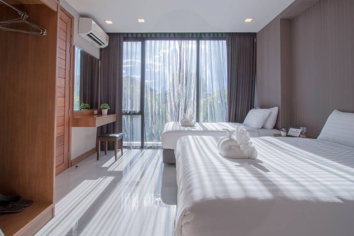 A hotel room with two beds, a wooden wardrobe, a small table with a plant, and a large window with curtains.