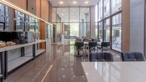 Una cafetería moderna con una barra larga, zona de mesas y grandes ventanales.